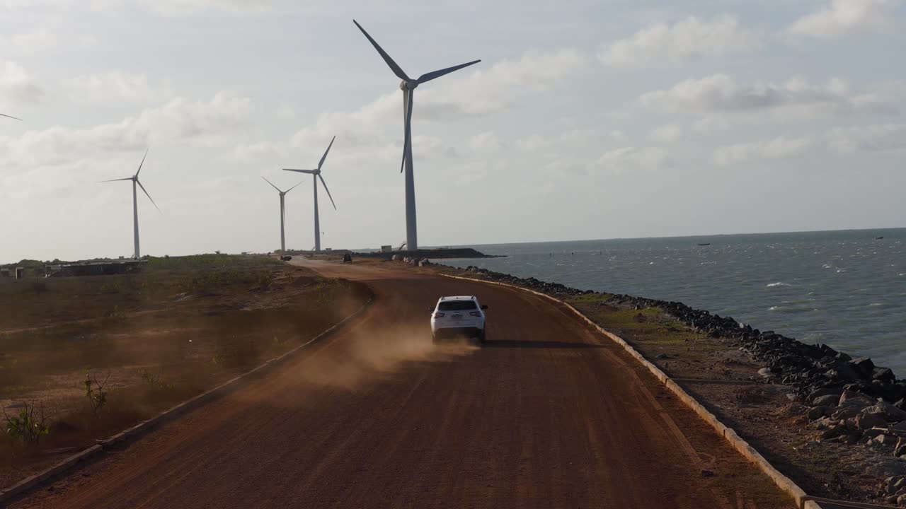 SUV che guida su una strada di servizio sterrata attraverso un parco eolico terrestre vicino a Ilha do Guajiru, Itarema, Ceará, Brasile, con turbine su scala industriale lungo l'argine costiero, ripresa con drone in movimento.