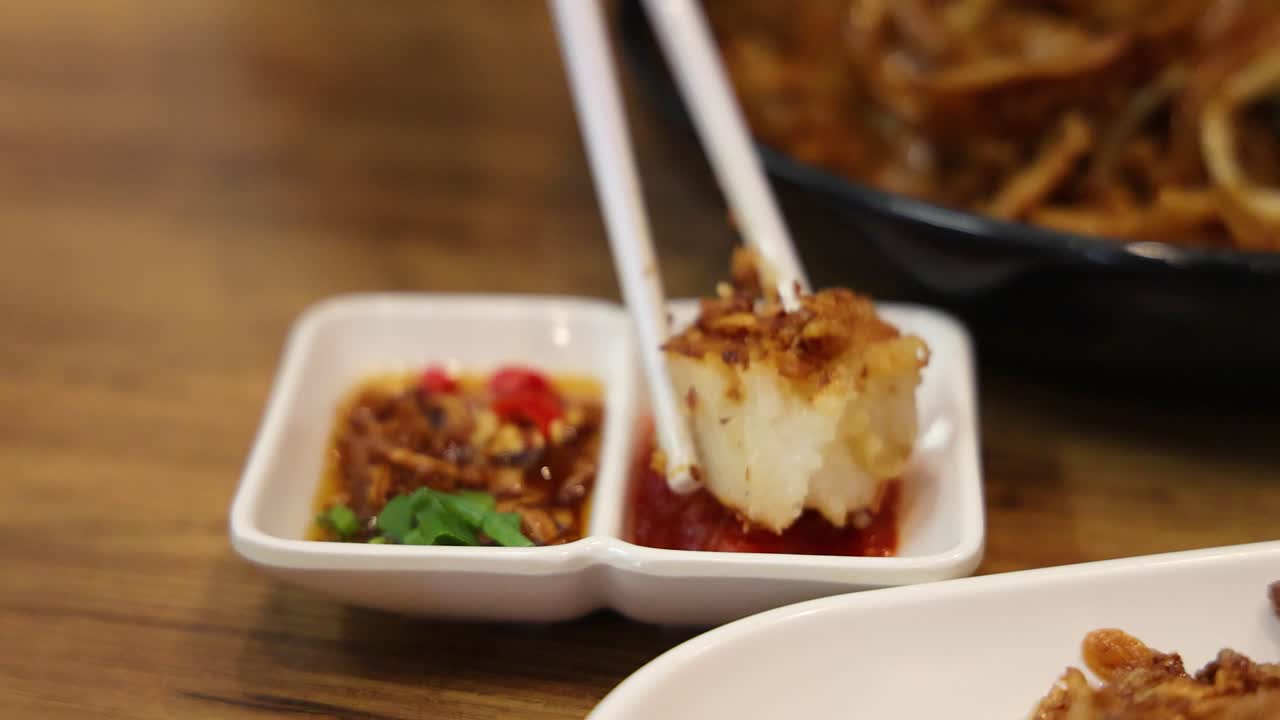 A piece of fried turnip cake is picked up with chopsticks and dipped into a spicy sauce in a restaurant setting with warm lighting