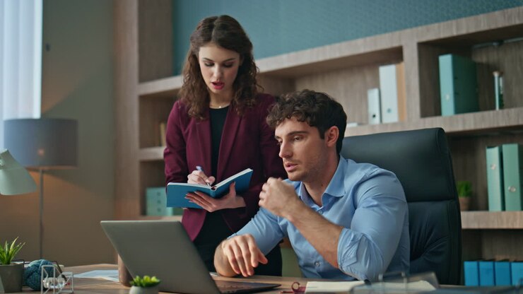 Manager giving instructions assistant making notes in copybook at office.