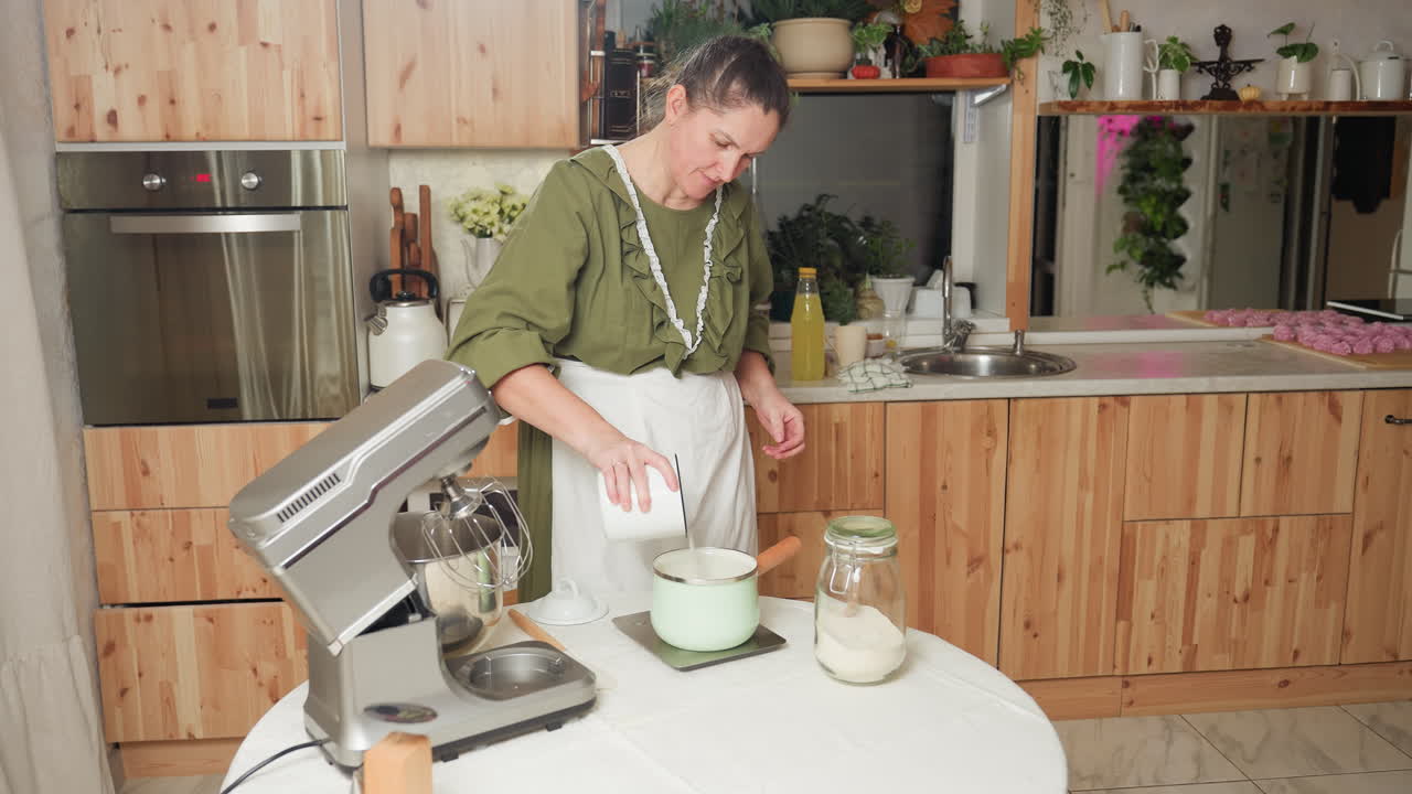 Baker in green dress and white apron pours flour from white container into mint green pot placed on kitchen scale, preparing baking ingredients beside mixer and jar in cozy home kitchen setup