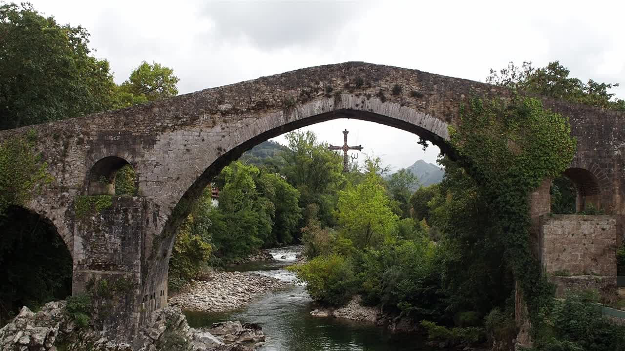 Static shot of the Roman bridge in Cangas de Onís, Asturias, Spain, with cross hanging under the arch and green forest around