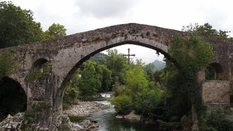 static shot of the roman bridge in cangas de onís, asturias, spain, with cross hanging under the arch and green forest around
