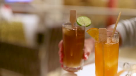 hand serving two colorful cocktails garnished with fruit slices slowly