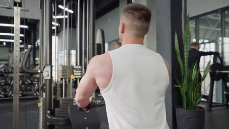 back view of fit male seated on bench pulling cable roller with controlled motion and focused effort against weight stack in busy gym environment showcasing strength training routine
