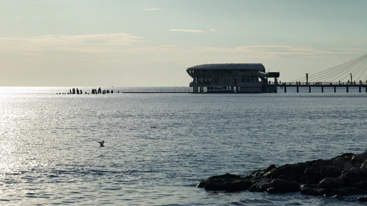 Sunlight reflects across the Adriatic Sea beside a pier in Durres, Albania. A waterfront building stands over the water beyond dark shoreline rocks.