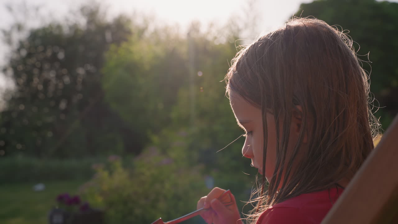 Girl Profile In Golden Hour Backyard, Soft Sunlight Outlining Face As She Inspects Small Object With Quiet Thoughtful Expression And Warm Natural Tones, Intimate Childhood Portrait