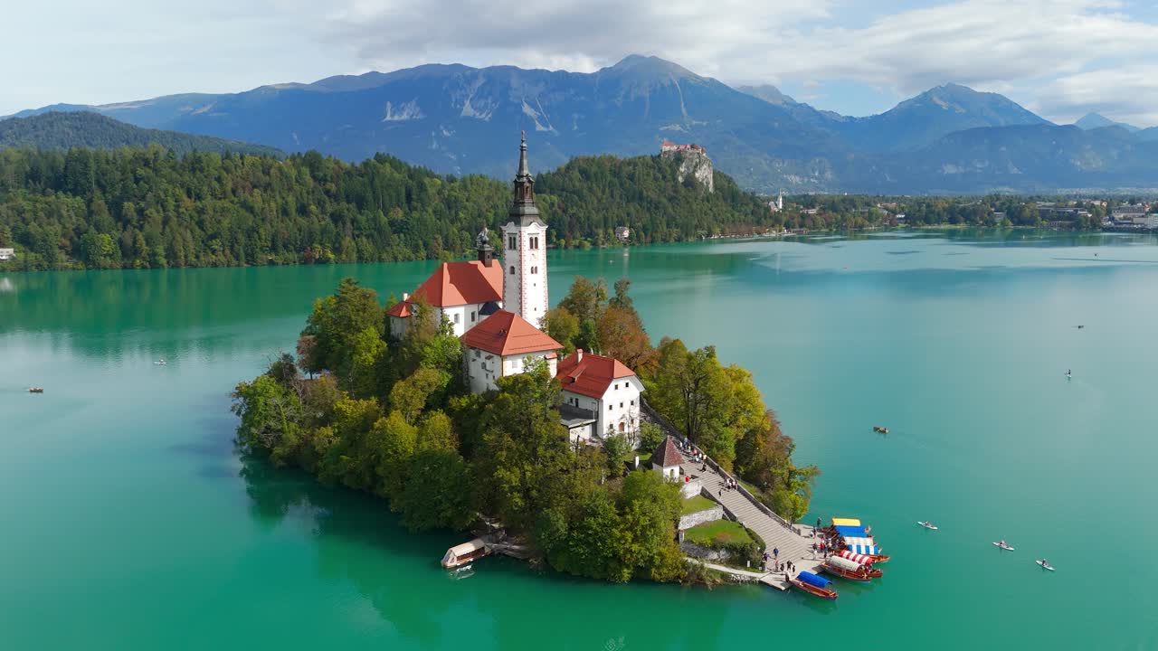 Ripresa aerea con drone che rivela l'Isola di Bled con la Chiesa dell'Assunzione, le acque turchesi del Lago di Bled, il Castello di Bled sulla scogliera e le Alpi Giulie sullo sfondo