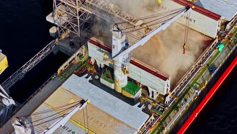 a dynamic top-down aerial parallax shot shows a crane and chute loading grain into the open hold of a large bulk carrier ship at the industrial port of riga, latvia