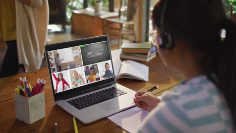 Schoolgirl-using-laptop-for-online-lesson-at-home,-with-diverse-teacher-and-class-on-screen