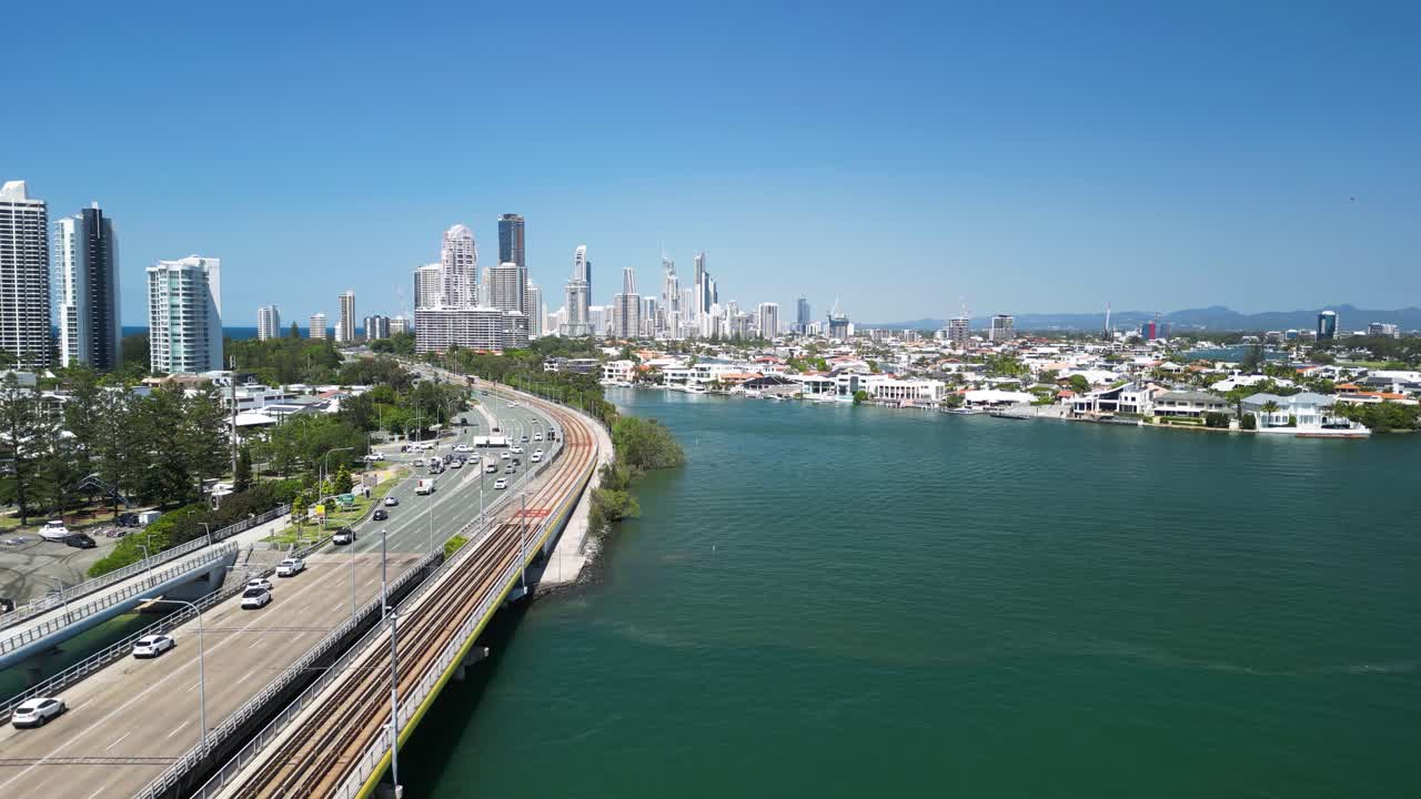 Modern electric tram travels away from a towering urban city skyline across a bridge over a coastal body of water. Gold Coast Queensland Australia
