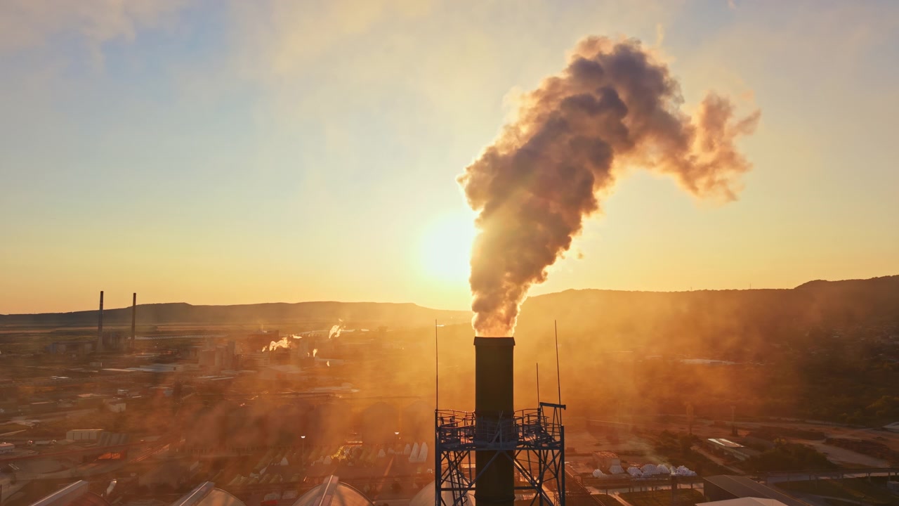 Il fumo sale da una ciminiera di fabbrica durante il tramonto in un'area industriale con colline e strutture distanti
