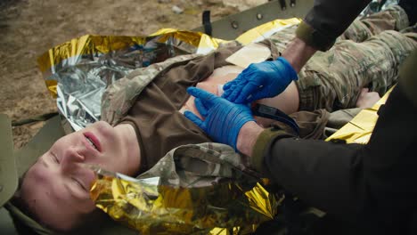 a confident male medic in blue medical gloves stops the blood of a wounded soldier in a camouflage uniform who is lying on a military medical stretcher in the combat area