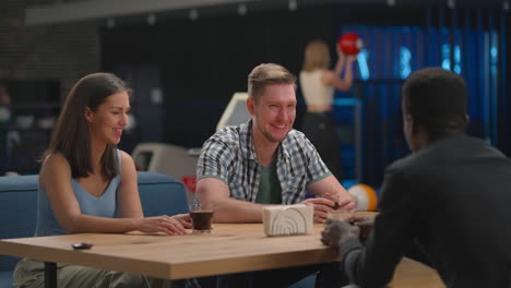 A-multi-ethnic-group-of-friends-sits-at-a-table-together-in-a-bowling-alley-and-talks.-Friends-met-together-in-a-bowling-alley-for-the-game