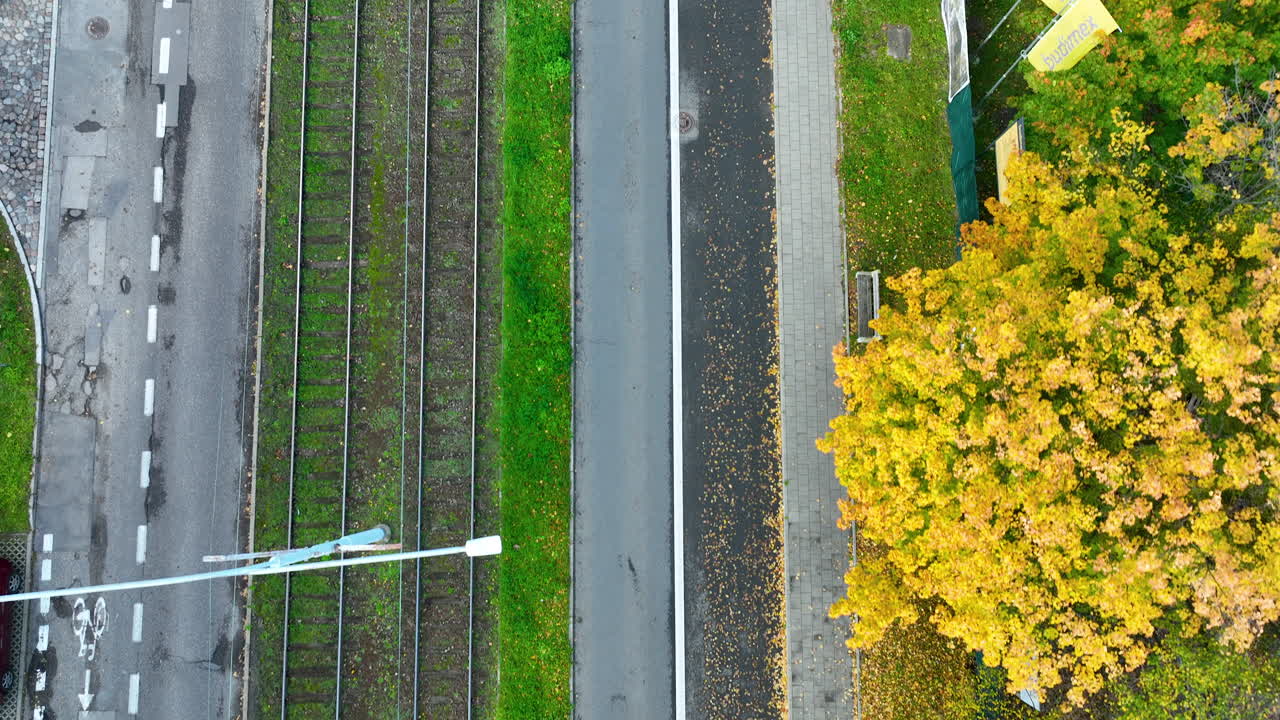 Aerial top-down view of a red tram moving along grass-covered tracks next to a bike lane and pedestrian path on a clear autumn day in Gdańsk, Poland