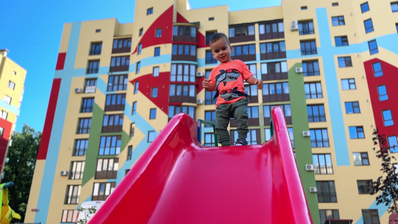 Beautiful baby boy climbs up the red slide outdoors. Cute toddler bangs his feet by the slide and then goes down. Colorful building at backdrop. Low angle view.