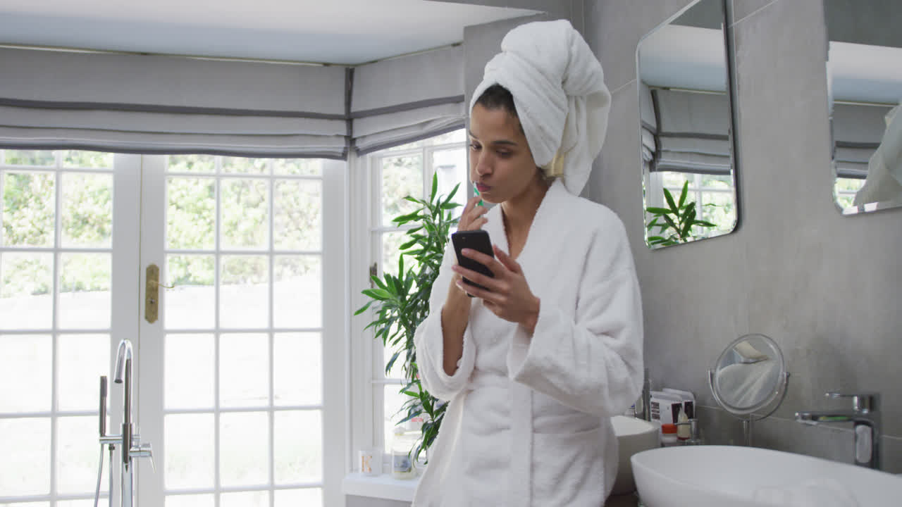 Mixed race woman brushing her teeth and using her smartphone in bathroom