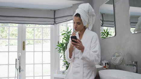 Mixed-race-woman-brushing-her-teeth-and-using-her-smartphone-in-bathroom