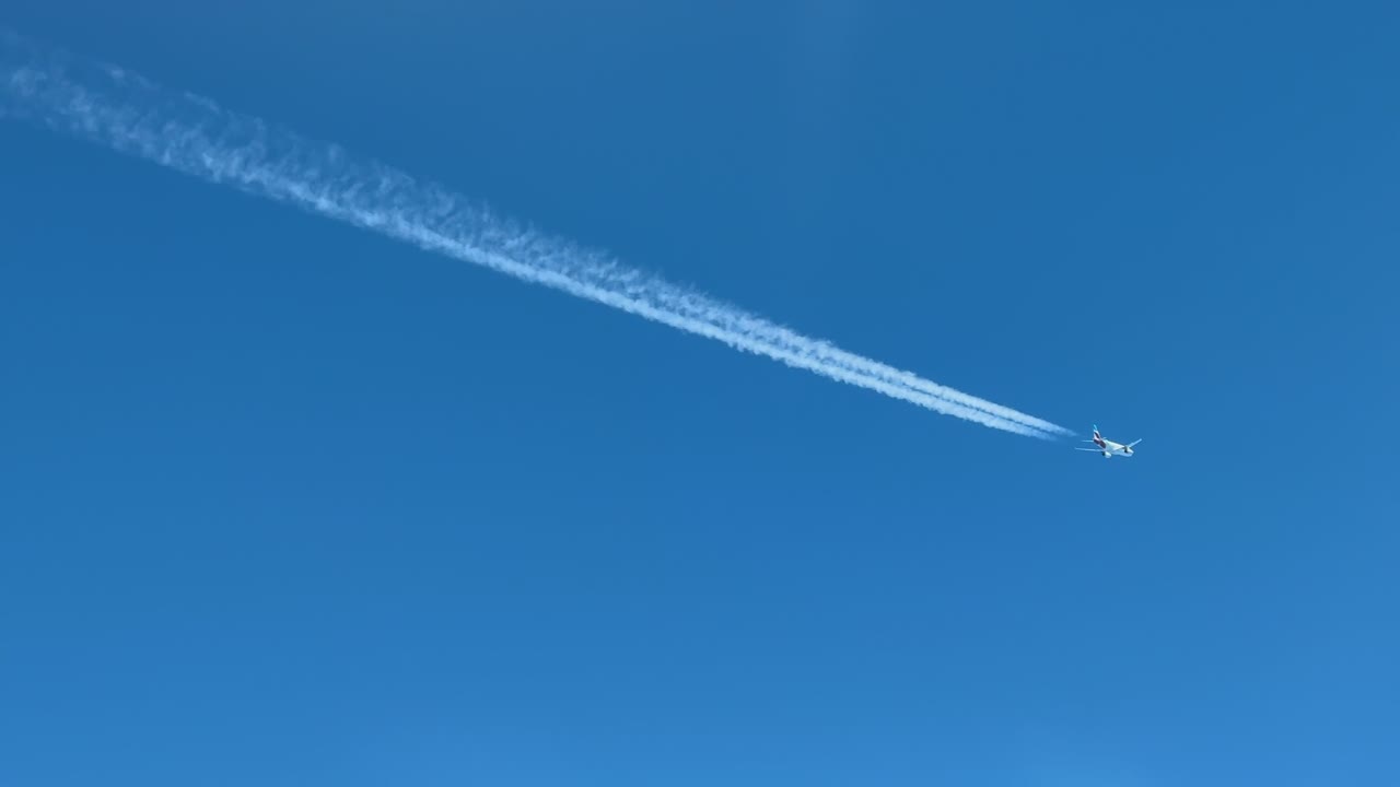 Aerial view of a twin-engine jet airplane flying at cruise leaving its contrail behind crossing diagonally a blue sky.