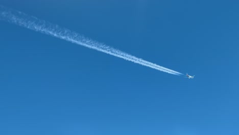 aerial view of a twin-engine jet airplane flying at cruise leaving its contrail behind crossing diagonally a blue sky.