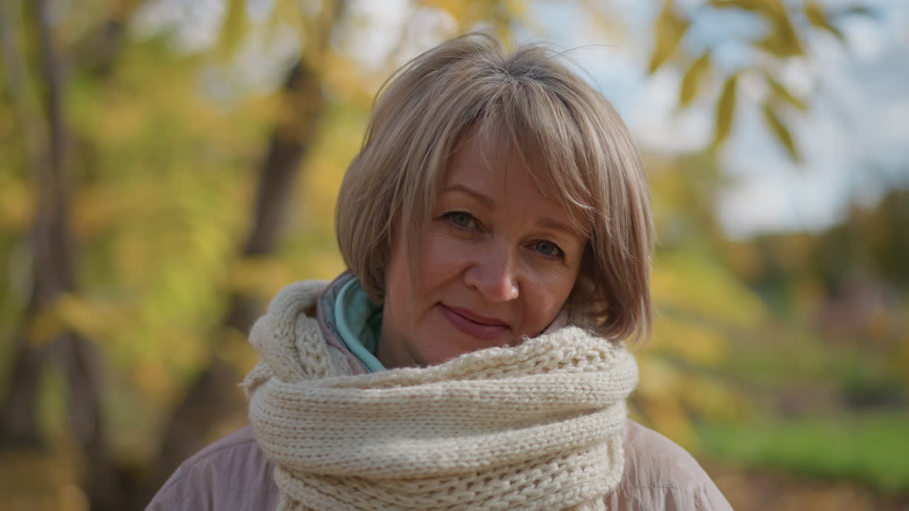 close up of woman in light jacket and thick scarf holding yellow leaf while smiling softly at camera in autumn park surrounded by golden foliage and trees under soft daylight atmosphere