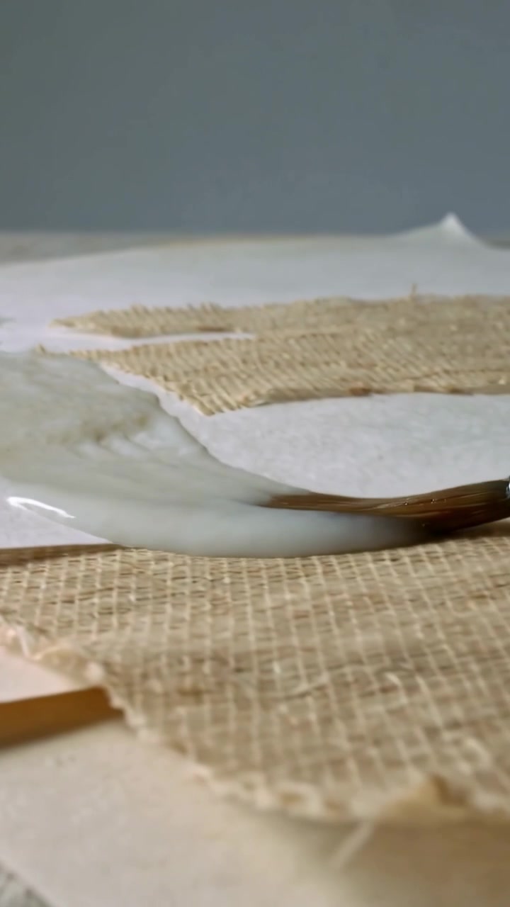 Vertical video: Pulling round brush from white pool, spreading medium at studio, priming burlap