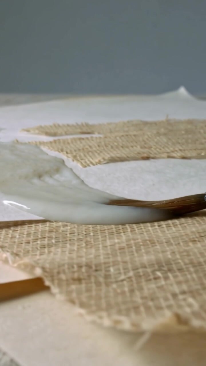 Vertical video: Pulling round brush from white pool, spreading medium at studio, priming burlap