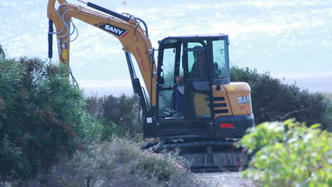 long shot of a plant machinery destroying semi-arid bushes near the beach on a sunny yet windy day, with the sparkling mediterranean sea in the background