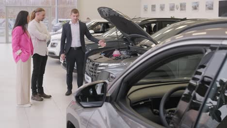 young car salesman showing to young couple new automobile at dealership salon.