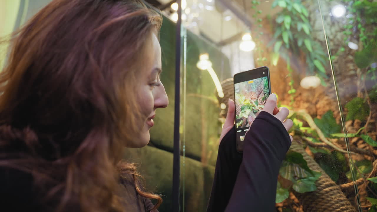 Side view of woman with pink acrylic nails using black phone to photograph vibrant greenery inside glass enclosure. Warm lighting and lush leaves reflect calm indoor nature experience