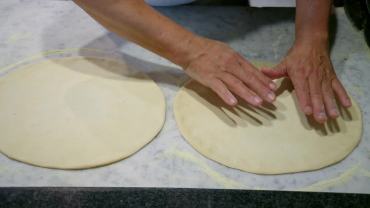 Hands shaping fresh pizza dough on a floured marble countertop, preparing for baking. Homemade cooking process
