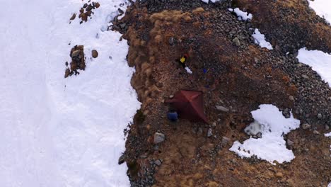 Man-next-camping-tent-over-rocky-area-on-snowy-mountain