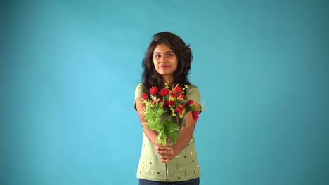 Una-Joven-India-Con-Una-Camiseta-Verde-Que-Muestra-Su-Ramo-De-Flores-Mirando-La-Cámara-Con-Una-Expresión-Tímida-Parada-En-Un-Estudio-Aislado-De-Fondo-Azul