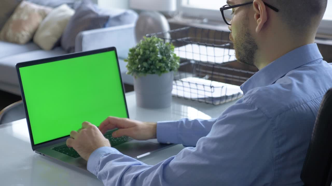 vista trasera de una pantalla de monitor verde y un joven trabajando en un proyecto de aprendizaje en línea en una computadora usando una computadora portátil en una habitación luminosa y acogedora en casa
