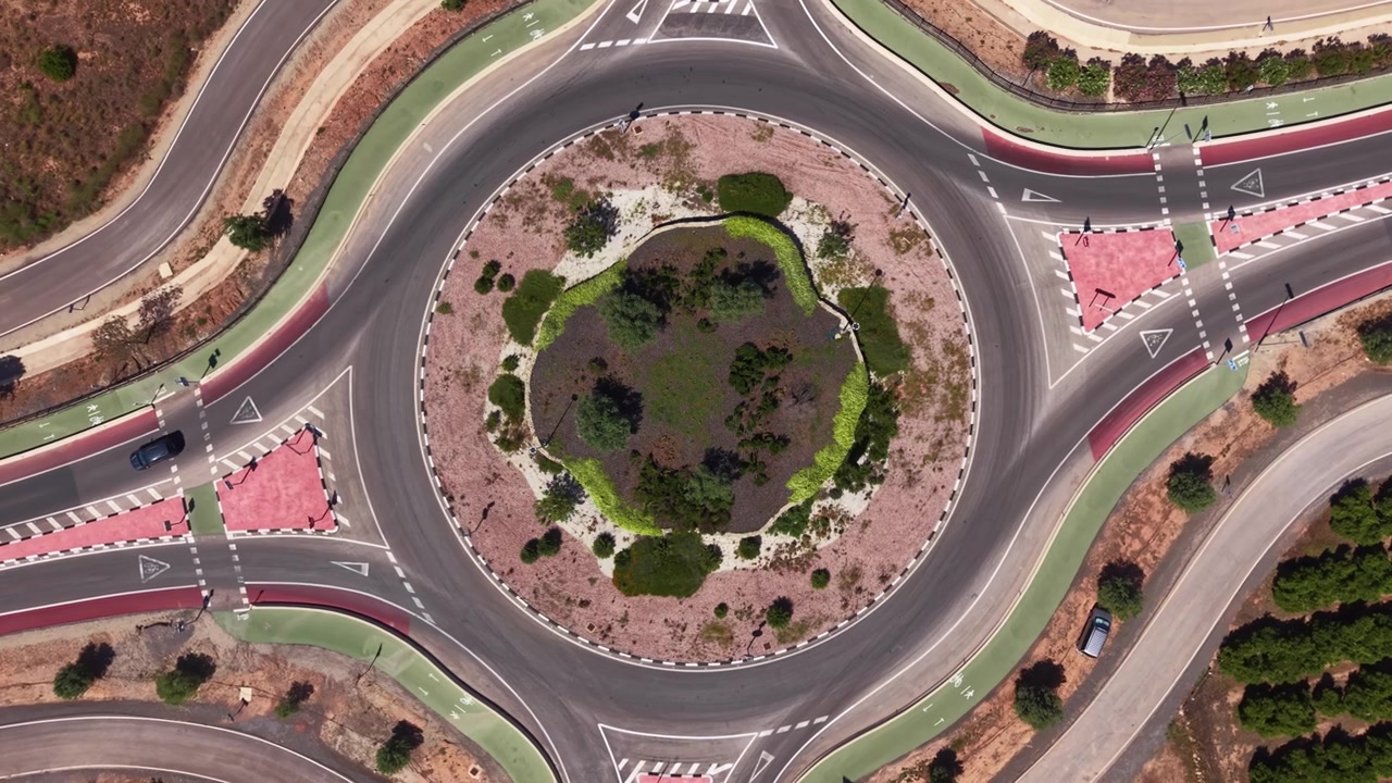 Vista dall'alto di una grande rotonda stradale con un'isola spartitraffico verde circolare e paesaggistica, attraversamenti pedonali rossi, segnaletica orizzontale bianca e traffico scarso