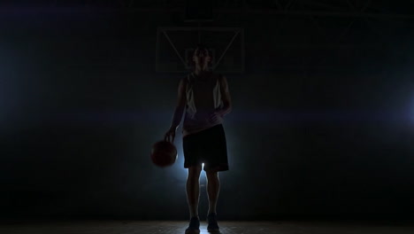 the basketball player stands on a dark playground and holds the ball in his hands and looks into the camera in the dark with a backlit in slow motion and around smoke