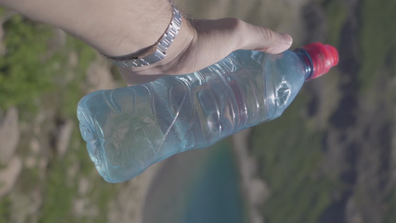 un hombre que sostiene una botella de agua en la mano junto a un lago y un bosque en las montañas en verano