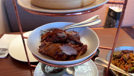 Traditional-crispy-peking-duck-with-steamed-pancakes,-onion,-cucumber,-hoisin-sauce-and-sriracha-sauce,-delicious-Asian-dish-served-on-a-three-floor-tray-tower,-4K-shot