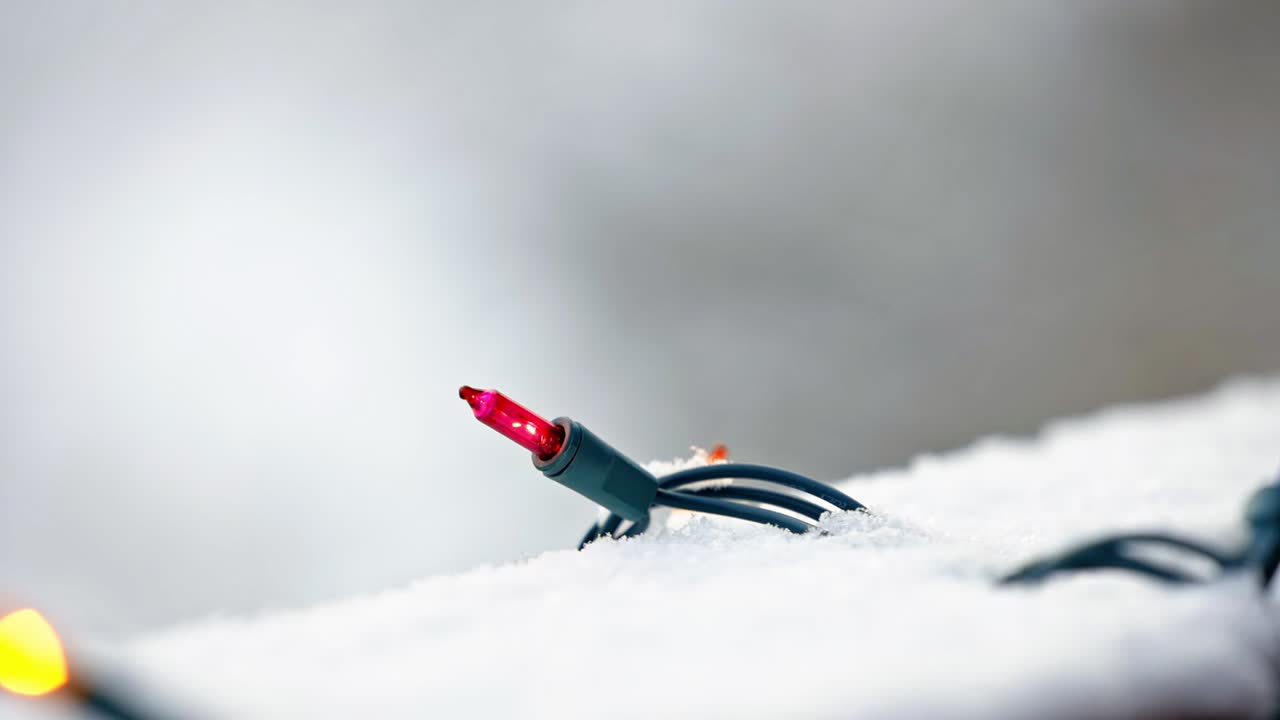 Close up of red and yellow Christmas lights on a snowy deck with a blurred background and snow falling