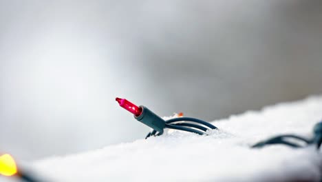 close up of red and yellow christmas lights on a snowy deck with a blurred background and snow falling