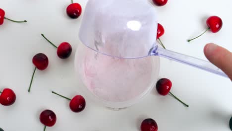 Vista-Estática-De-Pájaro-Desde-Una-Coctelera-Y-Una-Cuchara-Con-Un-Sabroso-Suero-De-Cereza-En-Polvo-Que-Se-Agrega-Con-Las-Cerezas-Circundantes-Sobre-Una-Base-Blanca