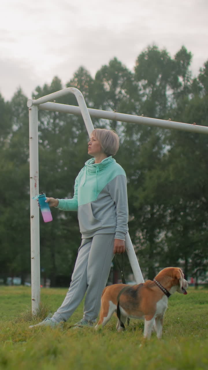 Mujer blanca con un beagle junto a una portería bebiendo de una botella reutilizable tras un paseo, con un aire de entrenador retirado, sudadera con capucha color menta, correa en la mano, campo de hierba y línea de árboles bajo un cielo nublado, ambiente de calma y compañía apacible.