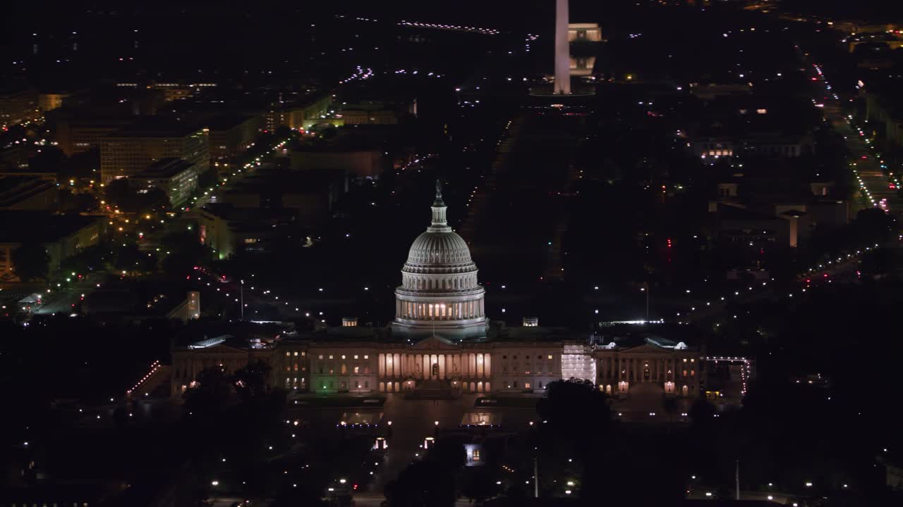 Tomada aérea noturna do Capitólio dos EUA iluminado, olhando para o National Mall em direção ao Monumento a Washington, em meio às luzes da cidade.