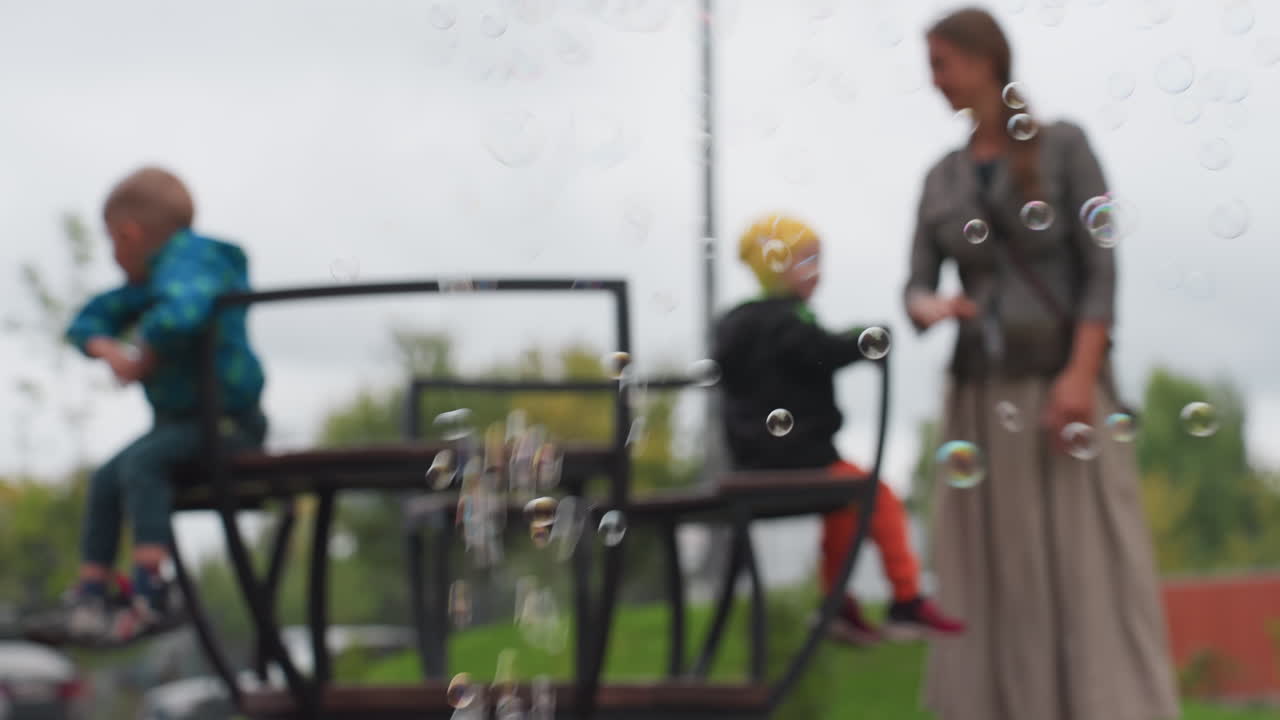 Blur background shows aunt spinning two kids on merry go round while soap bubbles drift across frame, soft focus seats and railings, cloudy sky over park, playful motion