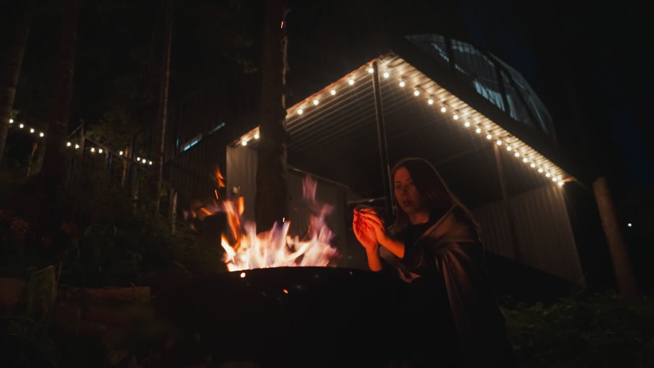 불에 의해 따뜻함을 얻기 위해 손으로 숨을 들이마시는 여자, 담요를 입은 아내, 숲 지역의 불빛에 캠프 하우스에 대항하여 서있다, 추운 저녁에 글램핑 사이트 안