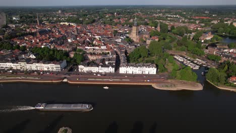 Medieval-city-seen-from-above-with-inland-shipping-large-cargo-vessel-leaving-ripple-waves-on-river-IJssel-passing-IJsselkade-boulevard-countenance-cityscape-of-tower-town-Zutphen