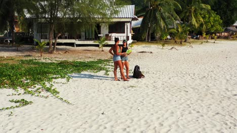 two girls with travel bags and tourist guide waiting on quiet exotic beach near resort cabins on tropical island in indonesia