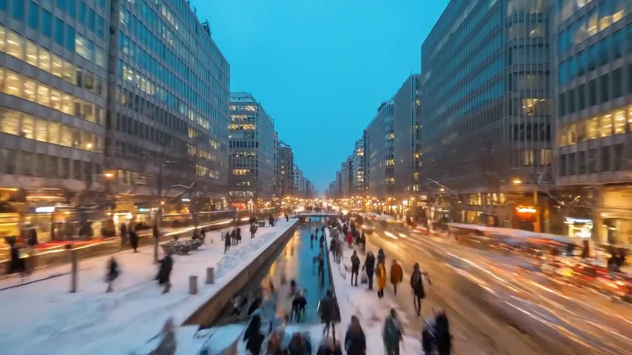 A bustling city street transitions from dusk to night, with glowing lights illuminating busy pedestrians and vehicles on a snowy winter evening.