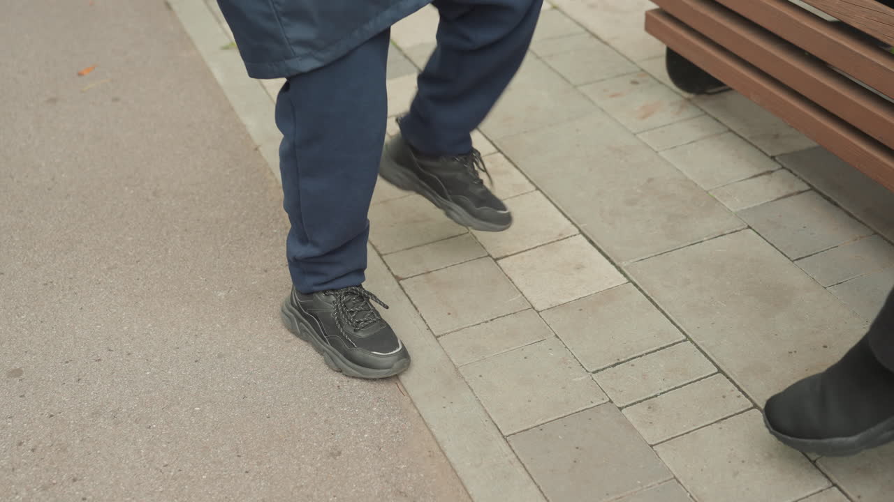 Legs and shoes of student standing near bench on paved sidewalk, casual sneakers with scuffed toes, coat hem visible, waiting, prepare for back to school, candid outdoor moment capturing footwear