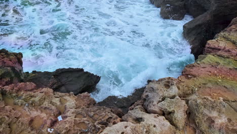 rocky coastal scene where turbulent white waves crash against jagged, reddish-brown rocks