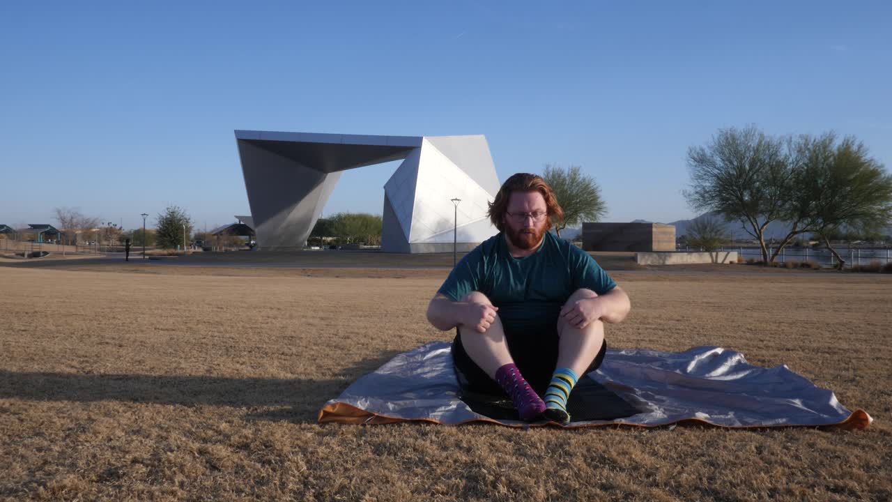 Red hair male doing sit ups in a park with an amphitheater behind them.